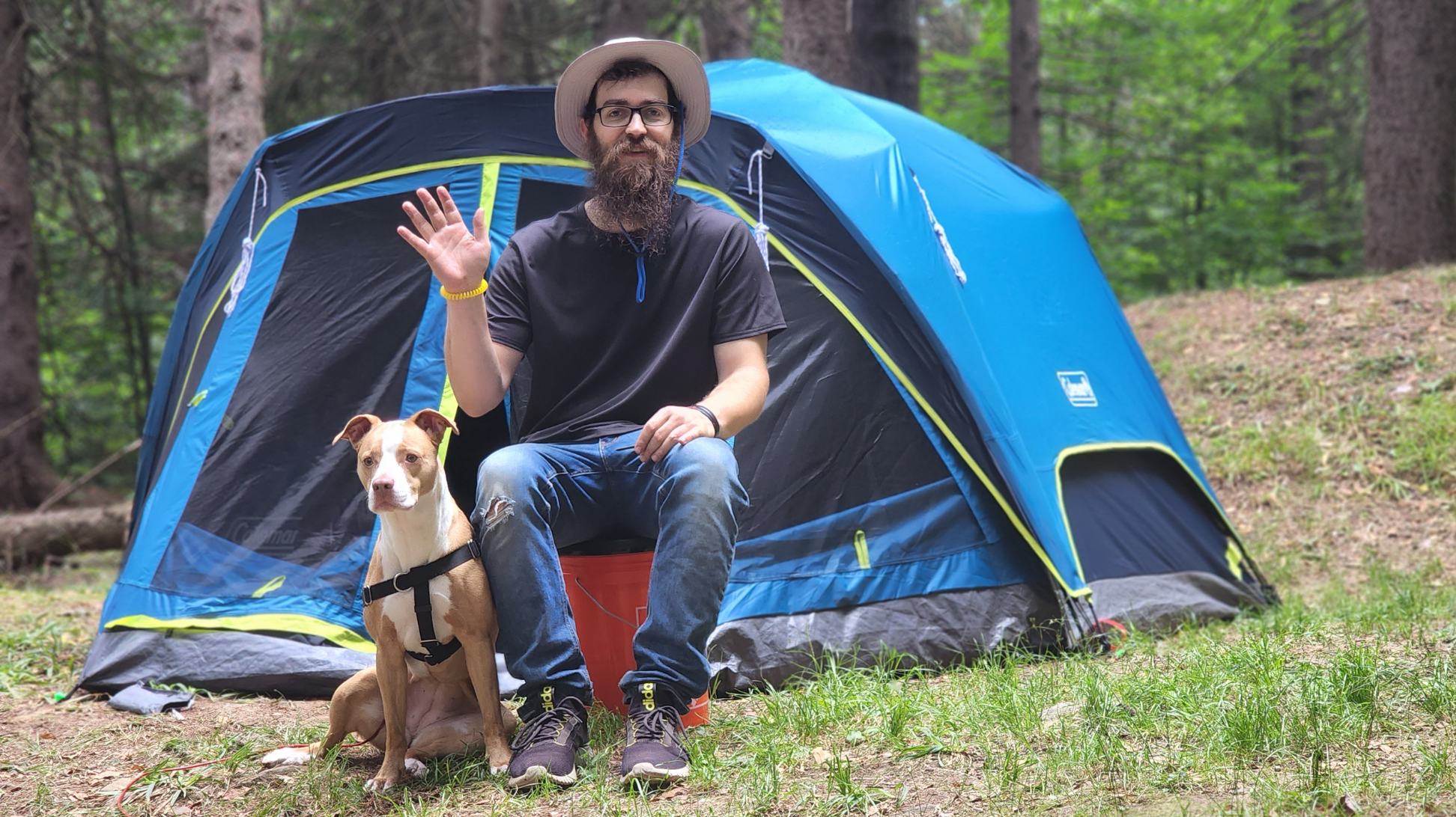 The image shows a bearded man wearing a hat sitting in front of a blue tent. The man is waving and holding the leash of his light brown and white pitbull boxer mix dog, also pictured.